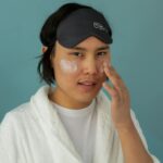 Jak krok po kroku wymienić cały zestaw kosmetyków na lepiej dobrany do męskiej skóry, nie wydając fortuny i nie podrażniając twarzy po drodze Azjata w szlafroku nakłada krem do twarzy, ma na czole opaskę do spania