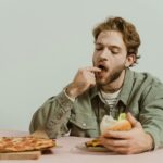 Mężczyzna w pomieszczeniu je pizzę i hamburgera, wygląda na zrelaksowanego